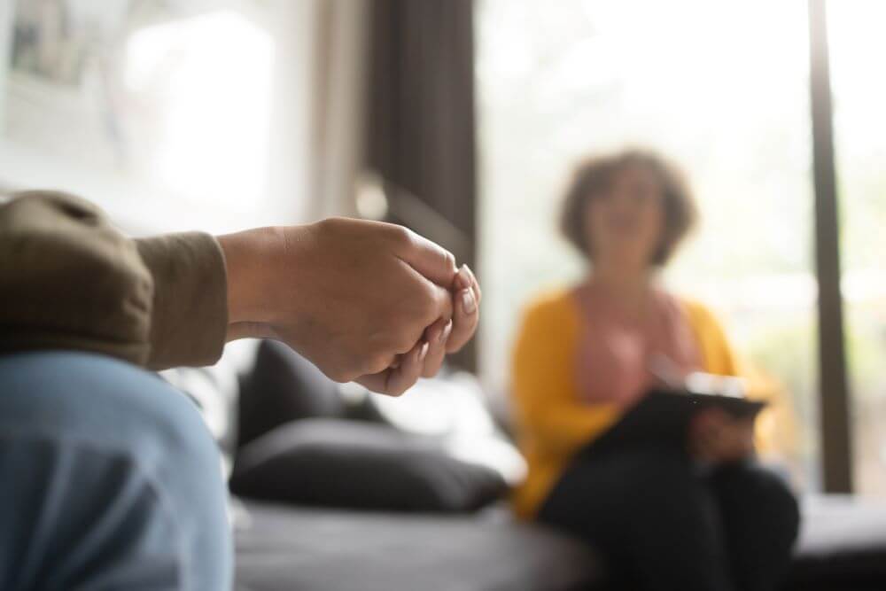 Photo of a person sitting with their hands clasped. A woman sits in the background looking on, out of focus.